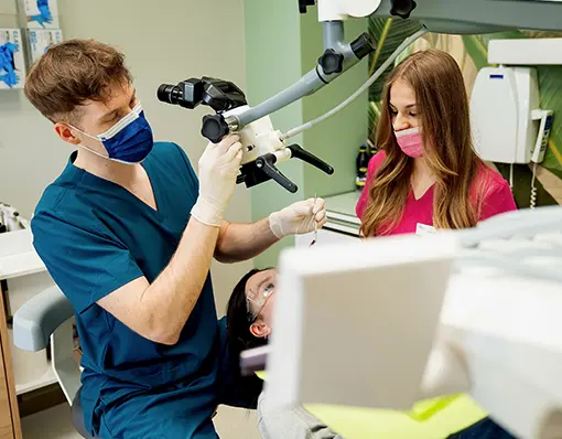 Lekarz stomatolog w ciemnoniebieskim fartuchu przeprowadza zabieg z użyciem mikroskopu endodontycznego, w asyście higienistki w różowym stroju medycznym.