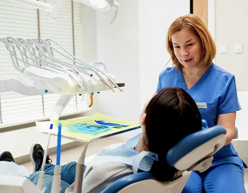 Lekarka stomatolog w niebieskim fartuchu medycznym rozmawia z pacjentką siedzącą na fotelu dentystycznym przed rozpoczęciem zabiegu.
