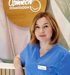Portret lekarki stomatolog w niebieskim fartuchu medycznym z identyfikatorem, na tle logo „Twój Uśmiech Stomatologia”.