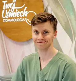Młody lekarz stomatolog w zielonym fartuchu medycznym, uśmiechający się na tle logo kliniki „Twój Uśmiech Stomatologia”.
