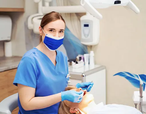 Lekarka stomatolog w niebieskim fartuchu i maseczce ochronnej bada zęby pacjentki leżącej na fotelu dentystycznym.