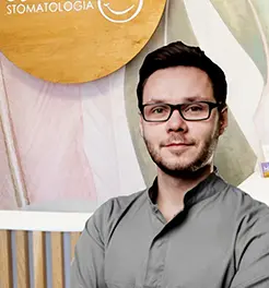 Lekarz stomatolog w okularach i szarym fartuchu medycznym na tle logo kliniki dentystycznej.