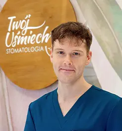 Młody lekarz stomatolog w ciemnoniebieskim fartuchu medycznym na tle drewnianego logo kliniki „Twój Uśmiech Stomatologia”.