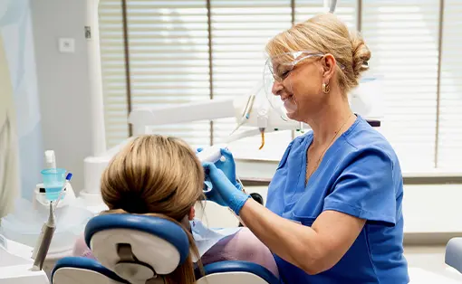 Lekarka stomatolog w niebieskim medycznym fartuchu i przyłbicy ochronnej bada zęby pacjentki siedzącej na fotelu dentystycznym.