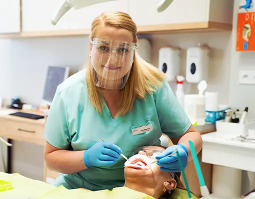 Dentystka w przyłbicy ochronnej i niebieskich rękawiczkach bada pacjentkę na fotelu stomatologicznym.