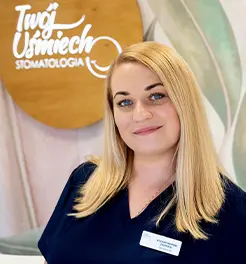 Uśmiechnięta kobieta w granatowym fartuchu medycznym na tle logo kliniki „Twój Uśmiech STOMATOLOGIA”.