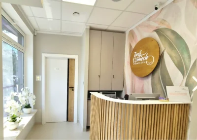 Recepcja kliniki stomatologicznej z drewnianą ladą i logo na ścianie.