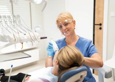 Dentystka w przyłbicy wykonująca badanie lub zabieg u pacjentki na fotelu stomatologicznym.